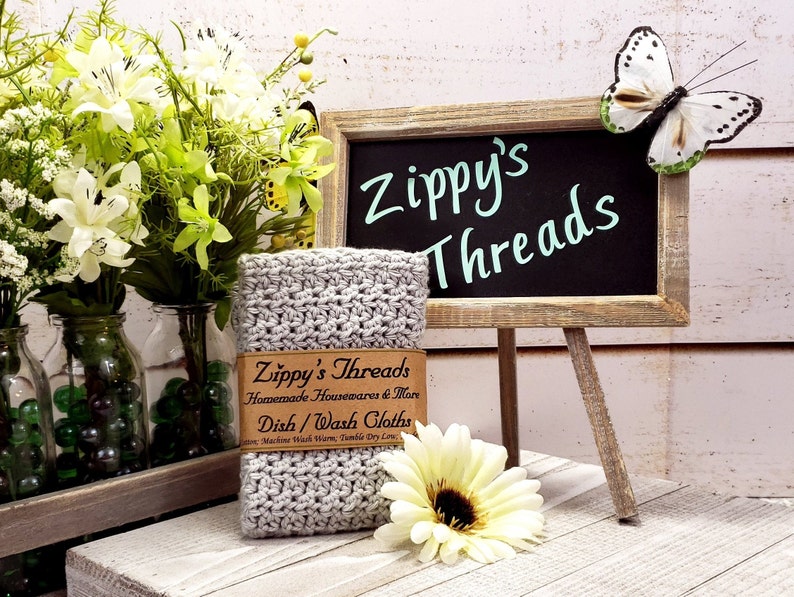 K&ouml;nnte beinhalten: Ein Stapel grauer geh&auml;kelter Geschirrt&uuml;cher mit einem braunen Etikett mit der Aufschrift "Zippy's Threads". Das Bild enth&auml;lt eine kleine Tafel mit den Worten "Zippy's Threads" und einen dekorativen Schmetterling. Ein Strau&szlig; wei&szlig;er und gr&uuml;ner Blumen befindet sich im Hintergrund.