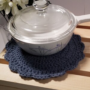 Pode incluir: Um descanso de panela azul de croch&ecirc; com borda ondulada, projetado para proteger as superf&iacute;cies do calor. Uma assadeira branca com tampa de vidro est&aacute; em cima. O prato tem um design floral e uma al&ccedil;a branca. O fundo inclui um copo e flores brancas.