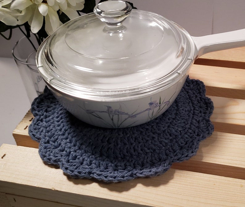 Pode incluir: Um descanso de panela azul de croch&ecirc; com borda ondulada, projetado para proteger as superf&iacute;cies do calor. Uma assadeira branca com tampa de vidro est&aacute; em cima. O prato tem um design floral e uma al&ccedil;a branca. O fundo inclui um copo e flores brancas.