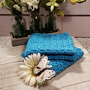 Puede incluir: Una pila de pa&ntilde;os de lavado de ganchillo de color turquesa brillante. Una margarita blanca y una mariposa decorativa con detalles en negro y verde descansan sobre los pa&ntilde;os. Jarrones de flores blancas y canicas verdes en el fondo.