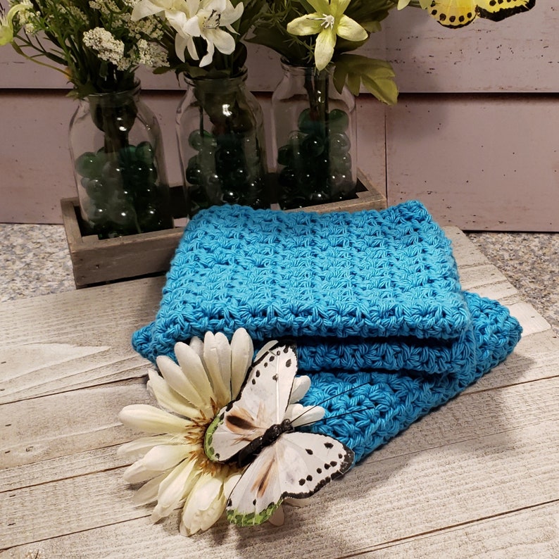 Puede incluir: Una pila de pa&ntilde;os de lavado de ganchillo de color turquesa brillante. Una margarita blanca y una mariposa decorativa con detalles en negro y verde descansan sobre los pa&ntilde;os. Jarrones de flores blancas y canicas verdes en el fondo.