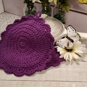 May include: Two purple crocheted pot holders with scalloped edges. The pot holders are on a wooden surface with a white butterfly and flower.