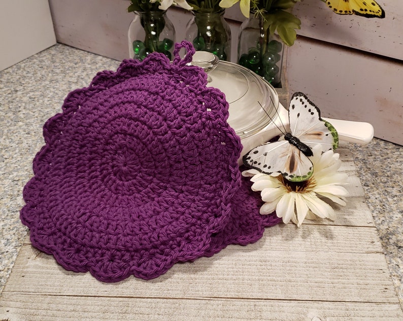 May include: Two purple crocheted pot holders with scalloped edges. The pot holders are on a wooden surface with a white butterfly and flower.