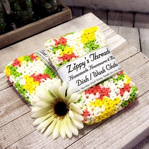 May include: Two crocheted dishcloths in a colourful pattern of green, yellow, white, and pink. The dishcloths are stacked on top of each other and have a label that reads "Zippy's Threads Homemade Housewares & More Dish/Wash Cloths Cotton. Machine Wash Warm. Tumble Dry Low. Made in the USA."
