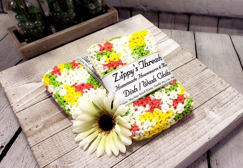 May include: Two crocheted dishcloths in a colourful pattern of green, yellow, white, and pink. The dishcloths are stacked on top of each other and have a label that reads "Zippy's Threads Homemade Housewares & More Dish/Wash Cloths Cotton. Machine Wash Warm. Tumble Dry Low. Made in the USA."