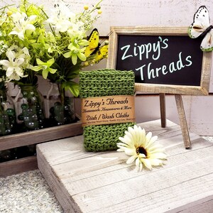 May include: Green knitted dishcloth with a brown label that reads "Zippy's Threads Handmade Housewares & More Dish/Wash Cloths Machine Wash Warm, Tumble Dry Low" on a wooden stand with a chalkboard sign that reads "Zippy's Threads".