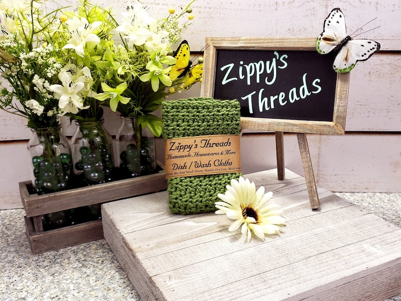 May include: Green knitted dishcloth with a brown label that reads "Zippy's Threads Handmade Housewares & More Dish/Wash Cloths Machine Wash Warm, Tumble Dry Low" on a wooden stand with a chalkboard sign that reads "Zippy's Threads".