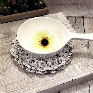 May include: A white ceramic bowl with a single yellow daisy inside, sitting on a gray crocheted coaster. The bowl has a handle and is on a light wood surface.