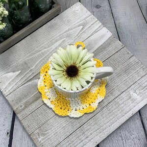 K&ouml;nnte beinhalten: Eine wei&szlig;e Keramiktasse auf einem gelb-wei&szlig; geh&auml;kelten Untersetzer. Eine gro&szlig;e, hellgr&uuml;ne und braune Blume befindet sich in der Tasse. Der Untersetzer hat einen gewellten Rand.