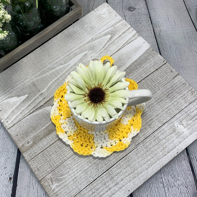 K&ouml;nnte beinhalten: Eine wei&szlig;e Keramiktasse auf einem gelb-wei&szlig; geh&auml;kelten Untersetzer. Eine gro&szlig;e, hellgr&uuml;ne und braune Blume befindet sich in der Tasse. Der Untersetzer hat einen gewellten Rand.