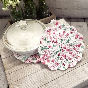 May include: A clear glass-lidded casserole dish rests on a white and floral patterned surface, accompanied by two handmade crochet pot holders. The pot holders feature a white base with pink and green accents.