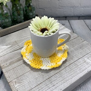 K&ouml;nnte beinhalten: Eine wei&szlig;e Keramiktasse auf einem gelb-wei&szlig; geh&auml;kelten Untersetzer. Die Tasse enth&auml;lt eine gro&szlig;e, hellgelbe Blume mit einem dunkelbraunen Zentrum. Der Untersetzer hat ein Schachbrettmuster und einen gewellten Rand.