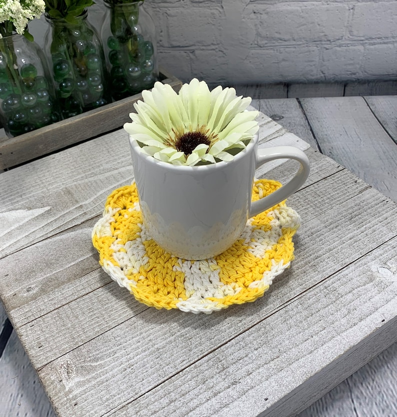 K&ouml;nnte beinhalten: Eine wei&szlig;e Keramiktasse auf einem gelb-wei&szlig; geh&auml;kelten Untersetzer. Die Tasse enth&auml;lt eine gro&szlig;e, hellgelbe Blume mit einem dunkelbraunen Zentrum. Der Untersetzer hat ein Schachbrettmuster und einen gewellten Rand.