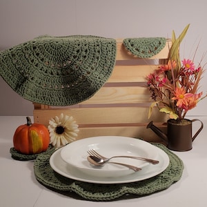 May include: A dining table setting featuring a green crocheted placemat, a white plate with silverware, and a matching crocheted doily. A small pumpkin, a white flower, and a bouquet of flowers in a watering can are also present.