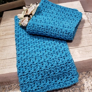 Puede incluir: Un conjunto de dos art&iacute;culos de ganchillo azul turquesa, posiblemente un pa&ntilde;o y una toalla, se exhiben sobre una superficie de madera r&uacute;stica. Una mariposa decorativa y una flor blanca a&ntilde;aden un toque delicado. El ganchillo presenta un patr&oacute;n de puntada texturizada.