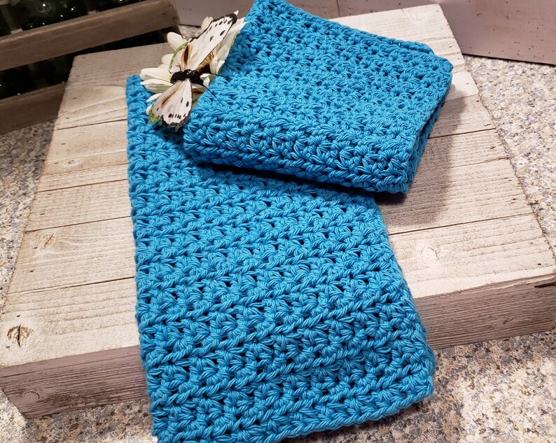 Puede incluir: Un conjunto de dos art&iacute;culos de ganchillo azul turquesa, posiblemente un pa&ntilde;o y una toalla, se exhiben sobre una superficie de madera r&uacute;stica. Una mariposa decorativa y una flor blanca a&ntilde;aden un toque delicado. El ganchillo presenta un patr&oacute;n de puntada texturizada.
