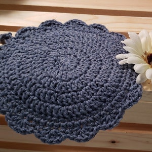 Pode incluir: Um pegador de panela redondo, feito &agrave; m&atilde;o, de croch&ecirc;, em uma cor azul-acinzentado suave. As bordas s&atilde;o onduladas, adicionando um toque decorativo. Uma flor branca e marrom &eacute; colocada &agrave; direita do pegador.