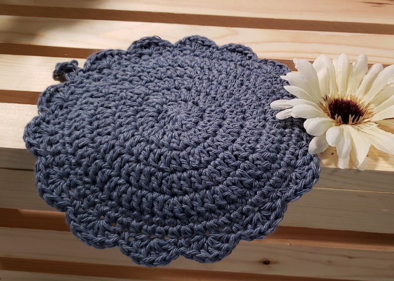 Pode incluir: Um pegador de panela redondo, feito &agrave; m&atilde;o, de croch&ecirc;, em uma cor azul-acinzentado suave. As bordas s&atilde;o onduladas, adicionando um toque decorativo. Uma flor branca e marrom &eacute; colocada &agrave; direita do pegador.