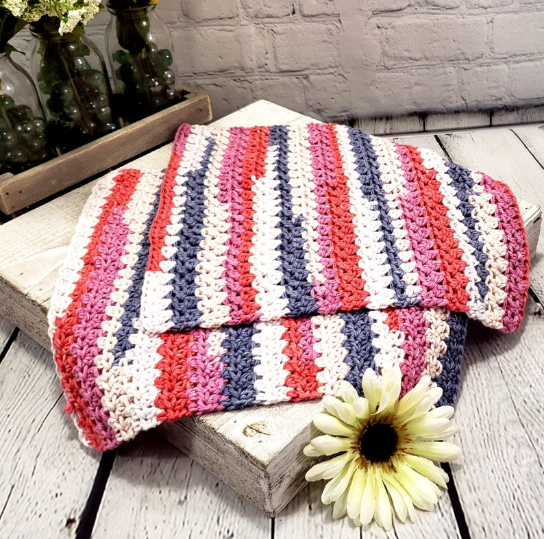 Pu&ograve; includere: Una coperta all'uncinetto a righe in tonalit&agrave; di rosa, bianco e grigio. La coperta &egrave; piegata e appoggiata su una superficie di legno bianco.