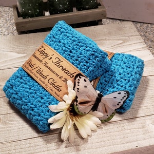 Puede incluir: Dos pa&ntilde;os de cocina turquesa tejidos a ganchillo est&aacute;n apilados, con una etiqueta marr&oacute;n que dice "Zippy's Threads Dish/Wash Cloths". Una mariposa blanca y una flor est&aacute;n colocadas encima. Los pa&ntilde;os de cocina est&aacute;n hechos a mano.