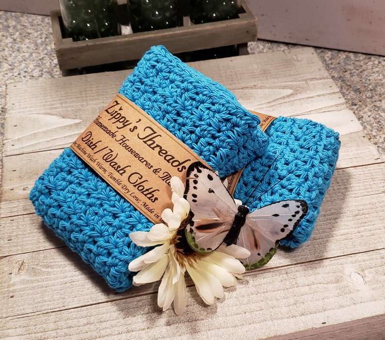 Puede incluir: Dos pa&ntilde;os de cocina turquesa tejidos a ganchillo est&aacute;n apilados, con una etiqueta marr&oacute;n que dice "Zippy's Threads Dish/Wash Cloths". Una mariposa blanca y una flor est&aacute;n colocadas encima. Los pa&ntilde;os de cocina est&aacute;n hechos a mano.