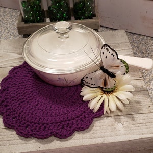 May include: A purple crocheted doily with a white and green butterfly on a white flower. A white ceramic covered dish with a floral design sits on the doily.