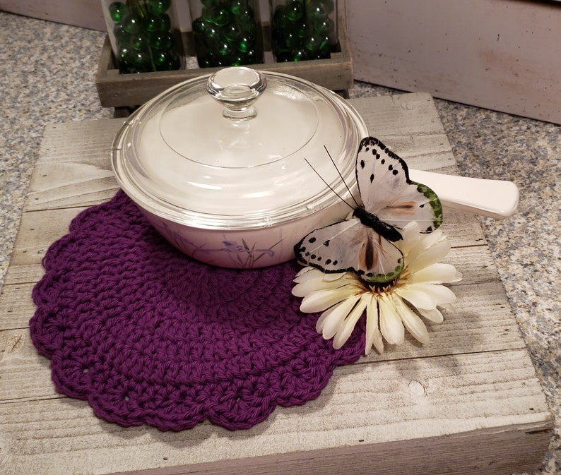 May include: A purple crocheted doily with a white and green butterfly on a white flower. A white ceramic covered dish with a floral design sits on the doily.