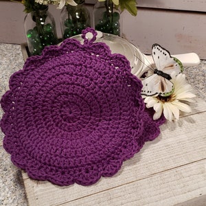 May include: Two purple crocheted pot holders with scalloped edges and a loop for hanging. The pot holders are on a white wooden surface.