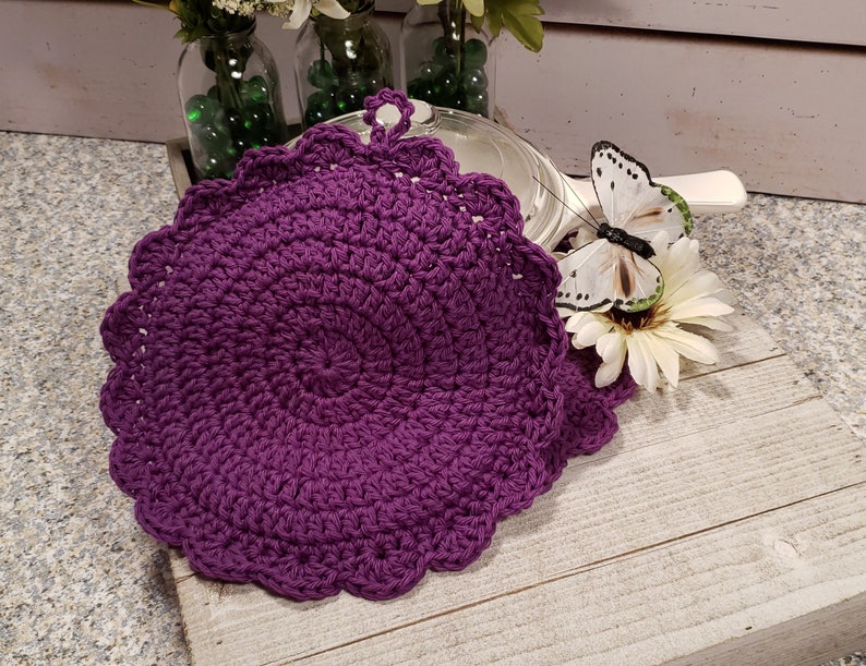 May include: Two purple crocheted pot holders with scalloped edges and a loop for hanging. The pot holders are on a white wooden surface.