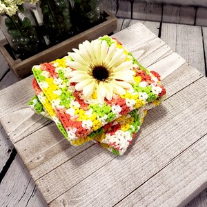May include: Three crocheted dishcloths stacked, featuring a yellow, green and pink colour pattern. A white flower with a yellow centre sits on top of the stack.