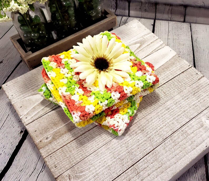 May include: Three crocheted dishcloths stacked, featuring a yellow, green and pink colour pattern. A white flower with a yellow centre sits on top of the stack.