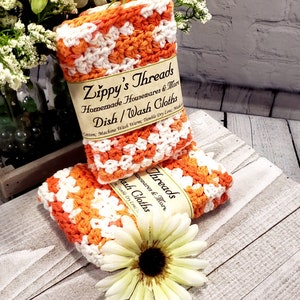 May include: Two orange and white crocheted dishcloths are stacked on a wooden surface. The dishcloths have a label that reads "Zippy's Threads Homemade Housewares & More Dish/Wash Cloths". A white and yellow flower is in the foreground.