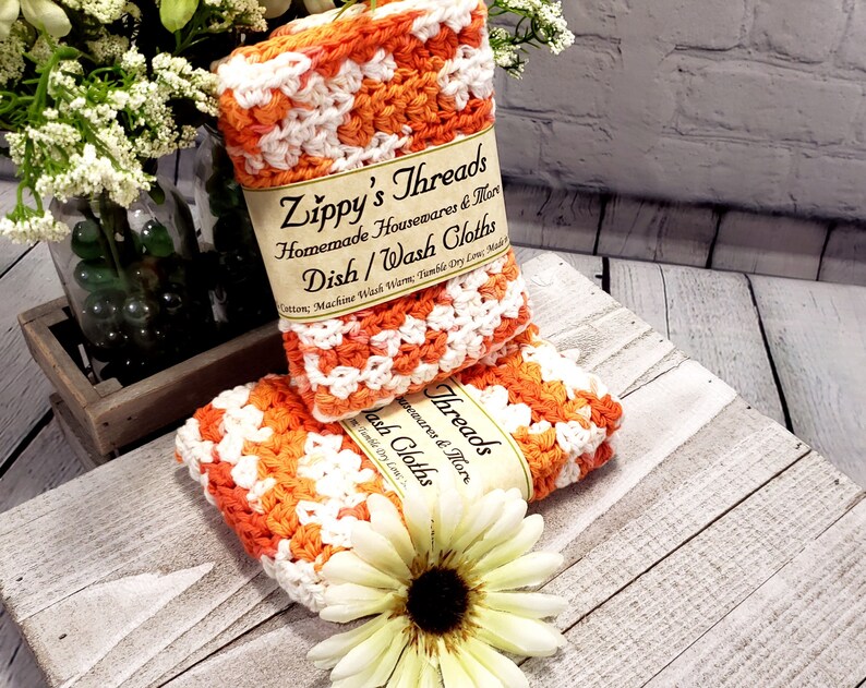 May include: Two orange and white crocheted dishcloths are stacked on a wooden surface. The dishcloths have a label that reads "Zippy's Threads Homemade Housewares & More Dish/Wash Cloths". A white and yellow flower is in the foreground.