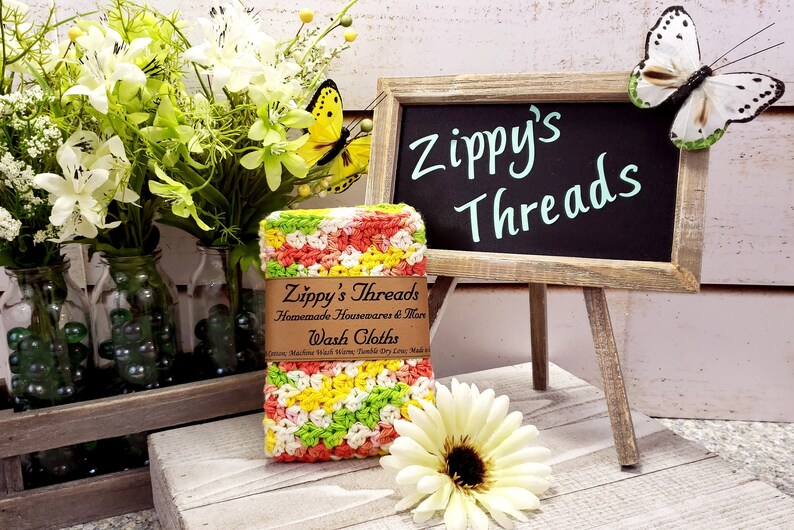 May include: A colourful crocheted washcloth with a label that reads "Zippy's Threads Homemade Housewares & More Wash Cloths".