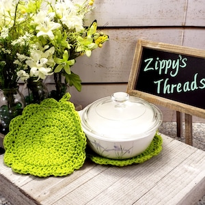 May include: Two green crocheted pot holders, one with a white ceramic casserole dish with a glass lid on top. The casserole dish is sitting on a wooden surface with a chalkboard sign that says "Zippy's Threads".