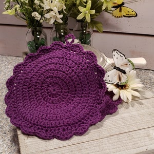 May include: Purple crocheted pot holder with a scalloped edge. The pot holder is on a white wooden surface with a white butterfly and flowers.