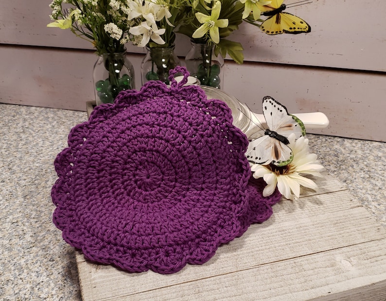 May include: Purple crocheted pot holder with a scalloped edge. The pot holder is on a white wooden surface with a white butterfly and flowers.