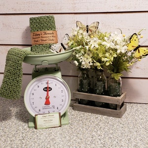 May include: A green kitchen scale with a white face and black numbers. The scale is sitting on a counter with a green crocheted dishcloth and a brown card with the text "Zippy's Threads Handmade Housewares & More Dish/Wash Cloths" on it. There is a bouquet of white flowers in glass jars in a wooden crate. The flowers are surrounded by butterflies, including two yellow butterflies.