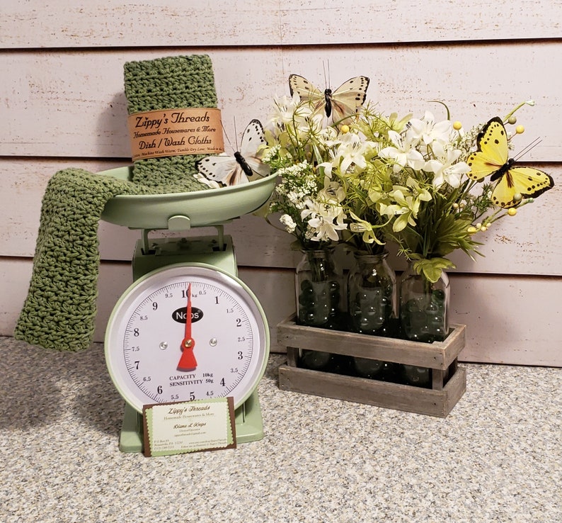May include: A green kitchen scale with a white face and black numbers. The scale is sitting on a counter with a green crocheted dishcloth and a brown card with the text "Zippy's Threads Handmade Housewares & More Dish/Wash Cloths" on it. There is a bouquet of white flowers in glass jars in a wooden crate. The flowers are surrounded by butterflies, including two yellow butterflies.
