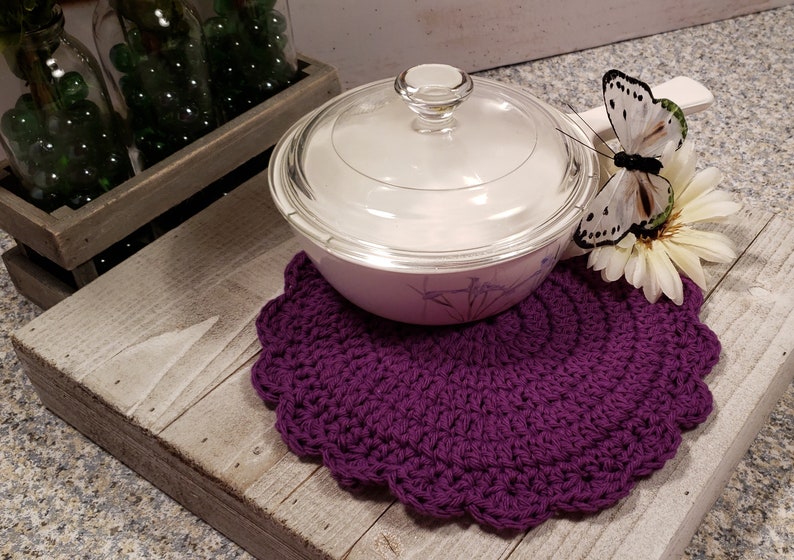 May include: Purple crochet pot holder with a white butterfly and a white flower on a wooden surface. A glass casserole dish with a lid is on top of the pot holder.