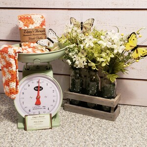 May include: A vintage-style mint green scale with a red needle displays orange and white washcloths from Zippy's Thread. Artificial flowers and butterflies decorate the scene. Three glass jars sit in a wooden crate, filled with flowers and butterflies.
