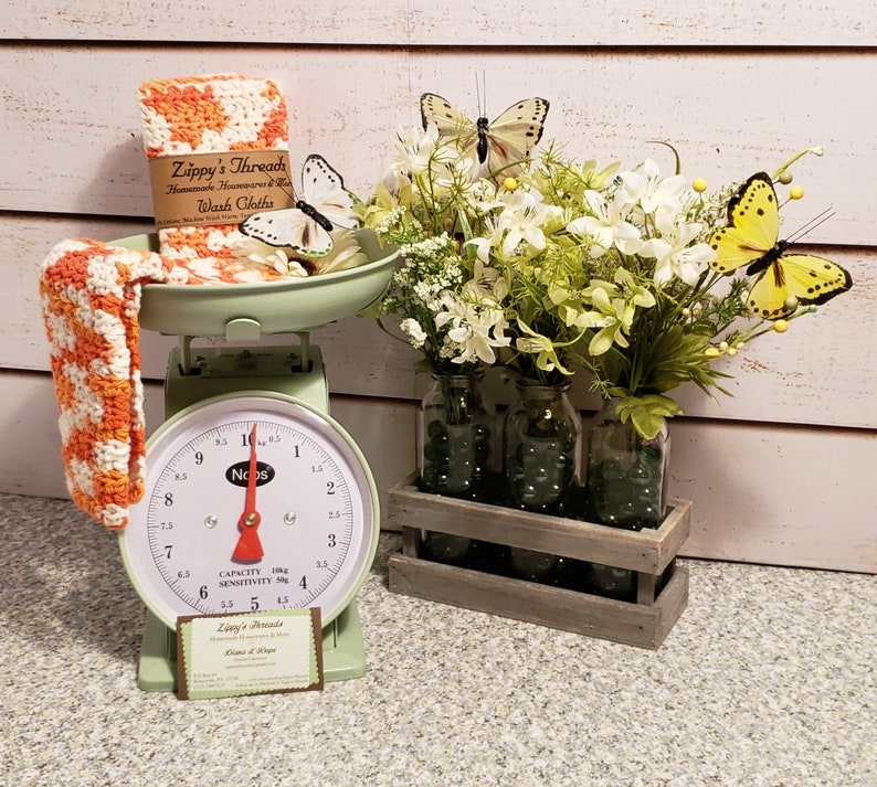 May include: A vintage-style mint green scale with a red needle displays orange and white washcloths from Zippy's Thread. Artificial flowers and butterflies decorate the scene. Three glass jars sit in a wooden crate, filled with flowers and butterflies.