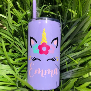 May include: Purple stainless steel tumbler with a unicorn design and the name "Emma" in pink lettering. The unicorn has a yellow horn, pink ears, and a flower crown with pink, blue, and green flowers. The tumbler comes with a clear plastic straw.