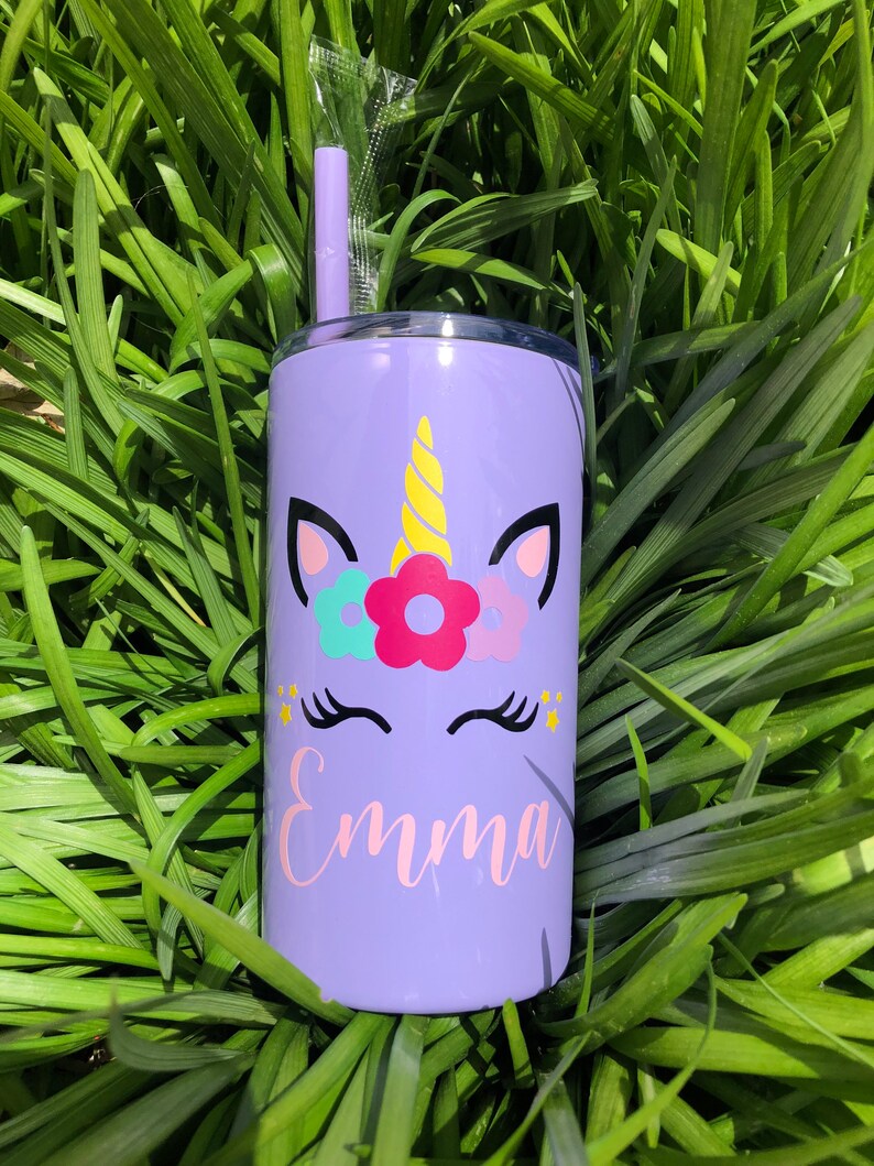 May include: Purple stainless steel tumbler with a unicorn design and the name "Emma" in pink lettering. The unicorn has a yellow horn, pink ears, and a flower crown with pink, blue, and green flowers. The tumbler comes with a clear plastic straw.