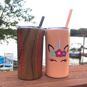 May include: Two stainless steel tumblers with lids and straws. One tumbler has a wood grain design with red baseball stitching. The other tumbler is peach colored with a unicorn face and flowers.