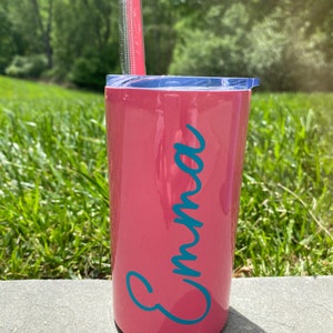 May include: A pink stainless steel tumbler with a blue lid and a teal blue name "Emma" on the side. A pink straw is in the tumbler.