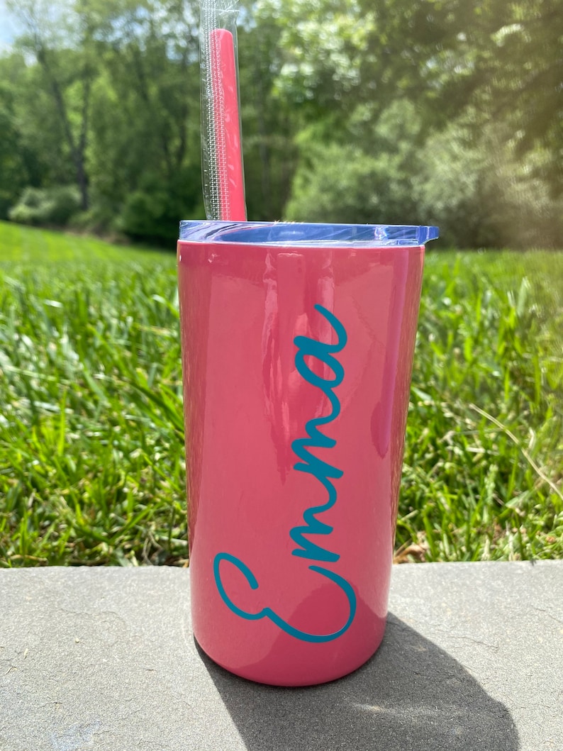 May include: A pink stainless steel tumbler with a blue lid and a teal blue name "Emma" on the side. A pink straw is in the tumbler.