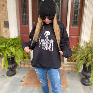 May include: Black sweatshirt with a white skeleton graphic wearing a peace sign. The skeleton is wearing a black beanie and sunglasses. The sweatshirt is being worn by a person with blonde hair and blue jeans.