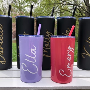 May include: Six personalized stainless steel tumblers with names "Danielle", "Ella", "Molly", "Emery", "Kelsey" and one other name that is unclear. The tumblers are in black, purple, and red.