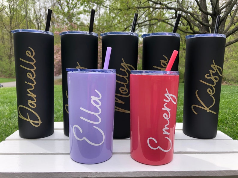 May include: Six personalized stainless steel tumblers with names "Danielle", "Ella", "Molly", "Emery", "Kelsey" and one other name that is unclear. The tumblers are in black, purple, and red.